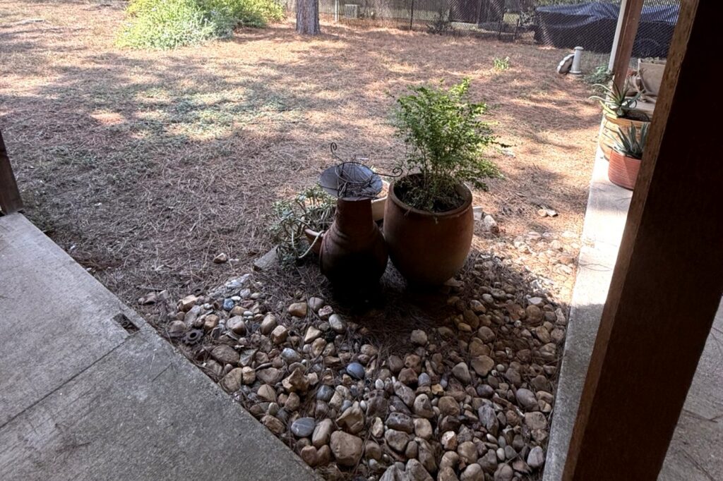 Pine needles built up in a river rock landscape bed against a house foundation in Bastrop, TX