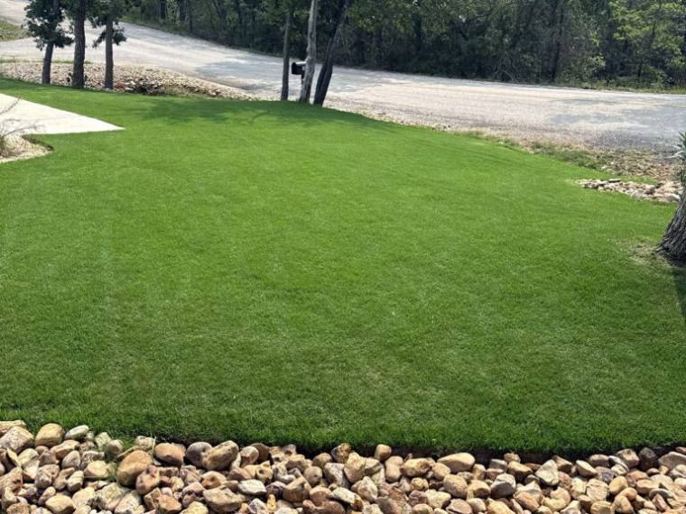 Manicured lawn edged with river rock beside a gravel drive under Bastrop oaks